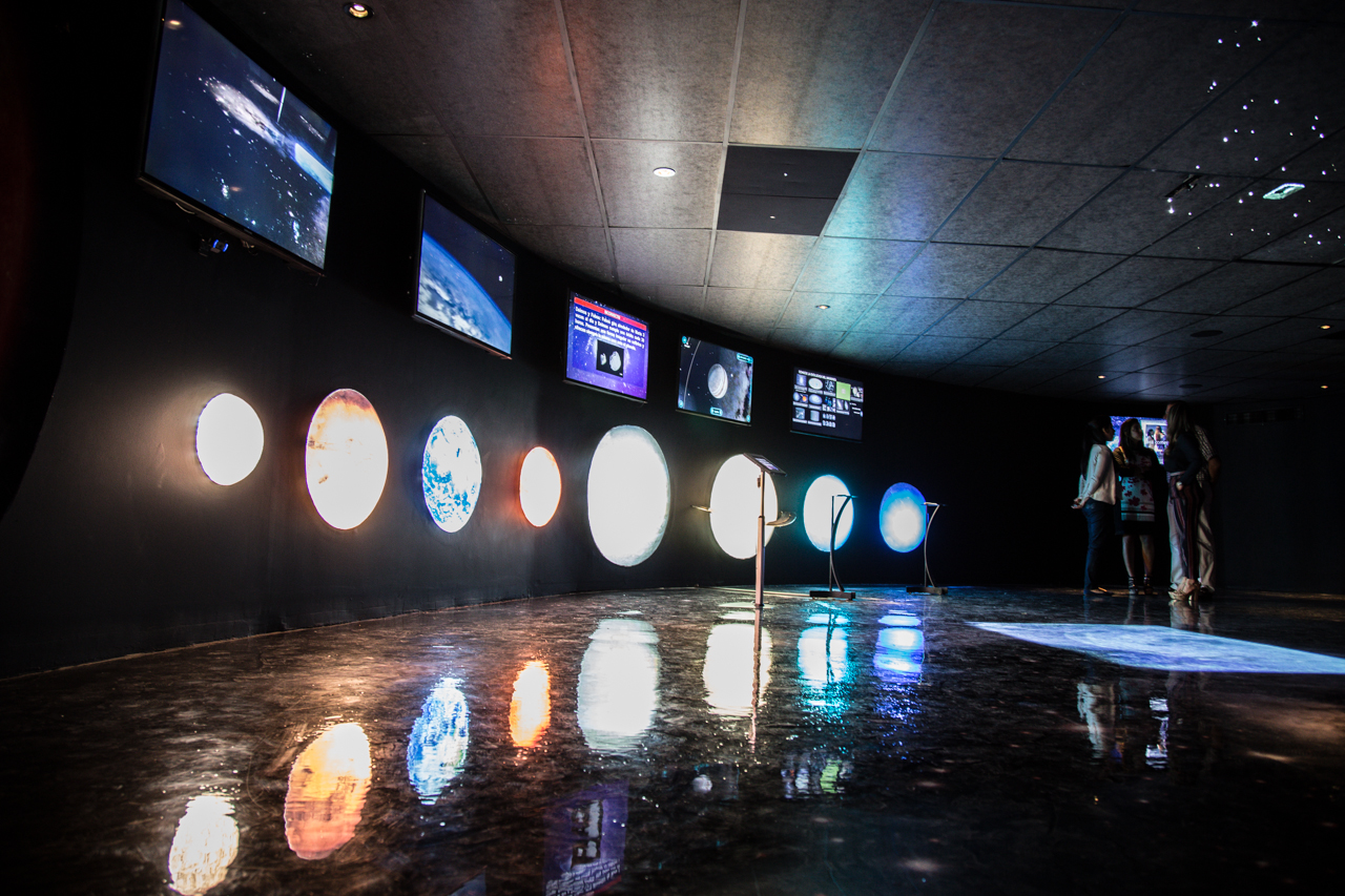 A modern audiovisual design exhibit featuring a series of illuminated circular displays reflecting on the glossy floor, with digital screens above showcasing various visual contents, aligning with the theme of ‘Emerging Future Trends in Audiovisual Design: VR, AR, and AI’. Una exposición moderna de diseño audiovisual que presenta una serie de pantallas circulares iluminadas reflejadas en el piso brillante, con pantallas digitales arriba mostrando varios contenidos visuales, alineados con el tema ‘Tendencias Futuras Emergentes en Diseño Audiovisual: VR, AR y AI’.