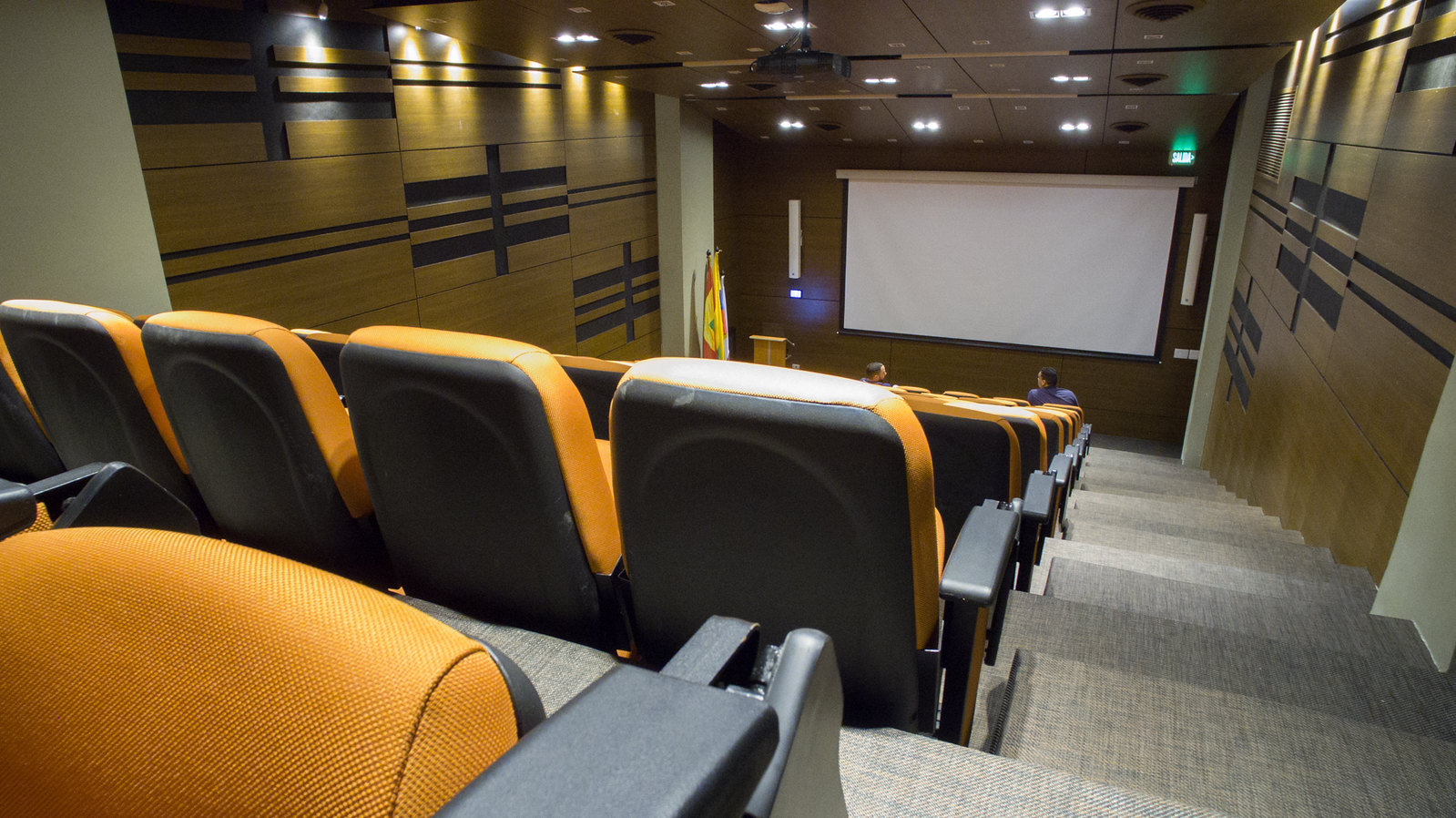 The Art of Theater Design: Stages, Spaces, and Audience Immersion 3 An interior view of a modern, well-lit auditorium with orange upholstered seats, decorative wooden panels, and a projection screen, optimized for an article on theater design. Vista interior de un auditorio moderno y bien iluminado con asientos tapizados en naranja, paneles de madera decorativos y una pantalla de proyección, optimizado para un artículo sobre diseño de teatros.