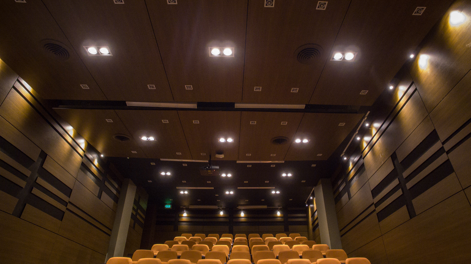 Mejorando las Experiencias de Audiovisual: El Papel Crucial del Diseño de Iluminación 3 Interior de un teatro moderno y bien iluminado con filas de asientos naranjas vacíos en orden ascendente, paredes de madera oscura con paneles acústicos integrados y un techo incrustado con múltiples luces redondas.