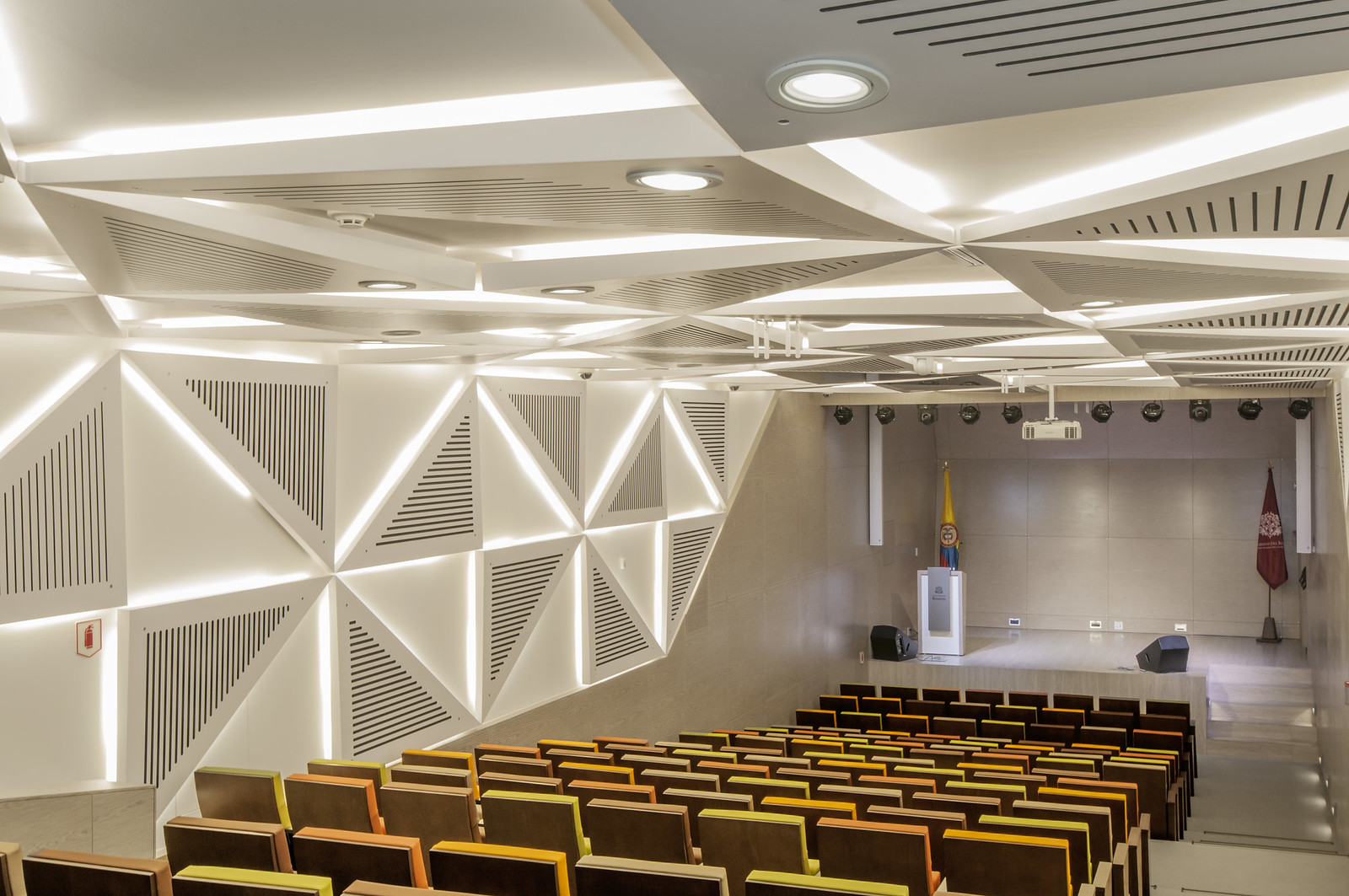 Interior view of a modern lecture hall with wooden chairs, geometric acoustic panels, and a podium, reflecting the commitment to sustainability in performing arts centers. Vista interior de un aula moderna con sillas de madera, paneles acústicos geométricos y un podio, reflejando el compromiso con la sostenibilidad en los centros de artes escénicas.