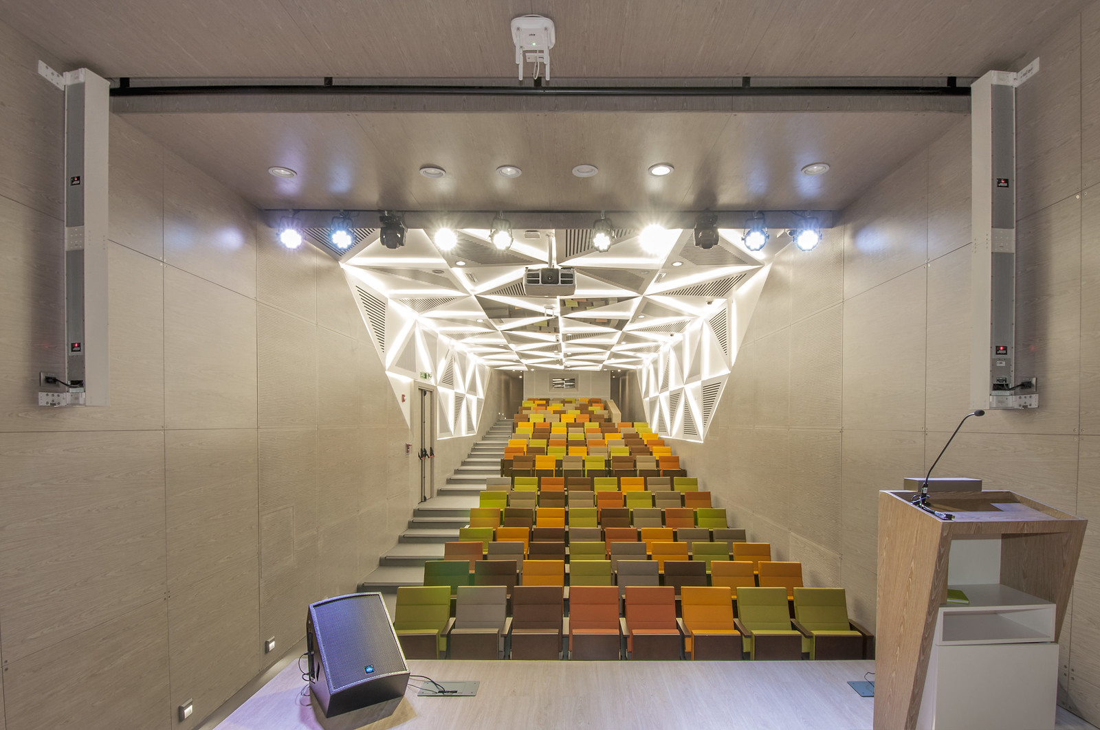 Empty modern auditorium with gradient colored seats and a wooden podium with a microphone, aligning with the article ‘Mastering Condenser Microphones: Placement, Accessories, and Care Tips’. Auditorio moderno vacío con asientos de colores en gradiente y un podio de madera con un micrófono, alineando con el artículo ‘Mastering Condenser Microphones: Placement, Accessories, and Care Tips’.
