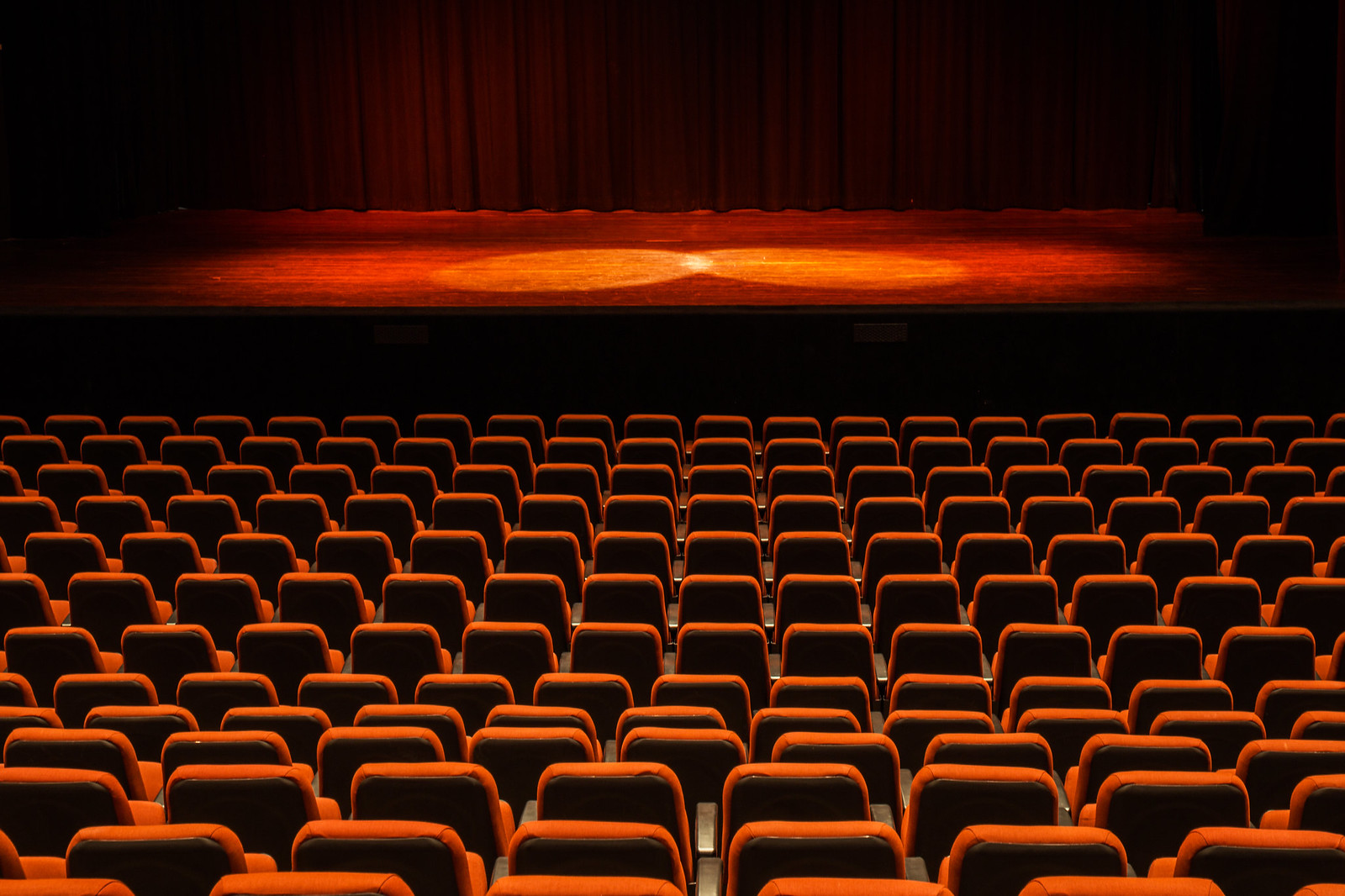 Interior view of a theater with a spotlight on the empty stage and rows of orange seats, reflecting the inclusive future of design trends in performing arts centers. Vista interior de un teatro con un foco de luz en el escenario vacío y filas de asientos naranjas, reflejando el futuro inclusivo de las tendencias de diseño en los centros de artes escénicas.