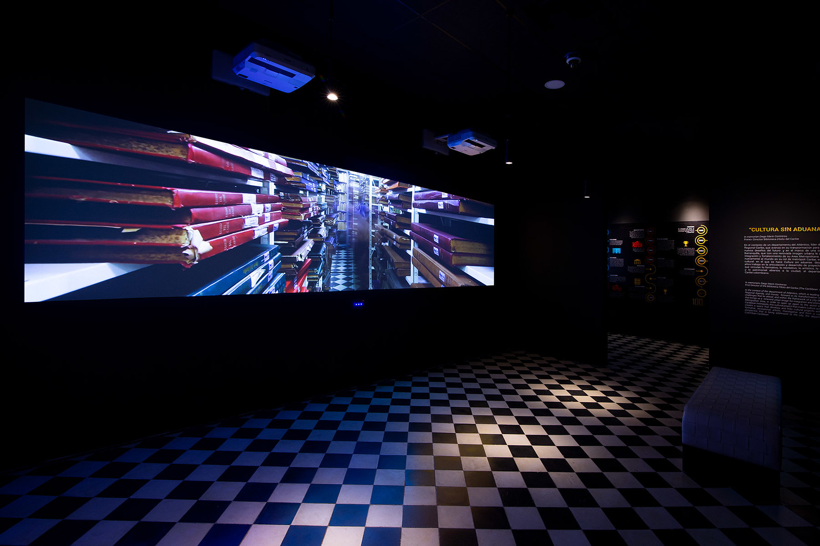 Innovaciones de Vanguardia en Pantallas y Sistemas de Proyección 1 Vista interior de una sala moderna con una gran pantalla que muestra una imagen abstracta y colorida, un banco blanco y un suelo a cuadros, relevante para el artículo ‘Innovaciones de vanguardia en pantallas y sistemas de proyección’.