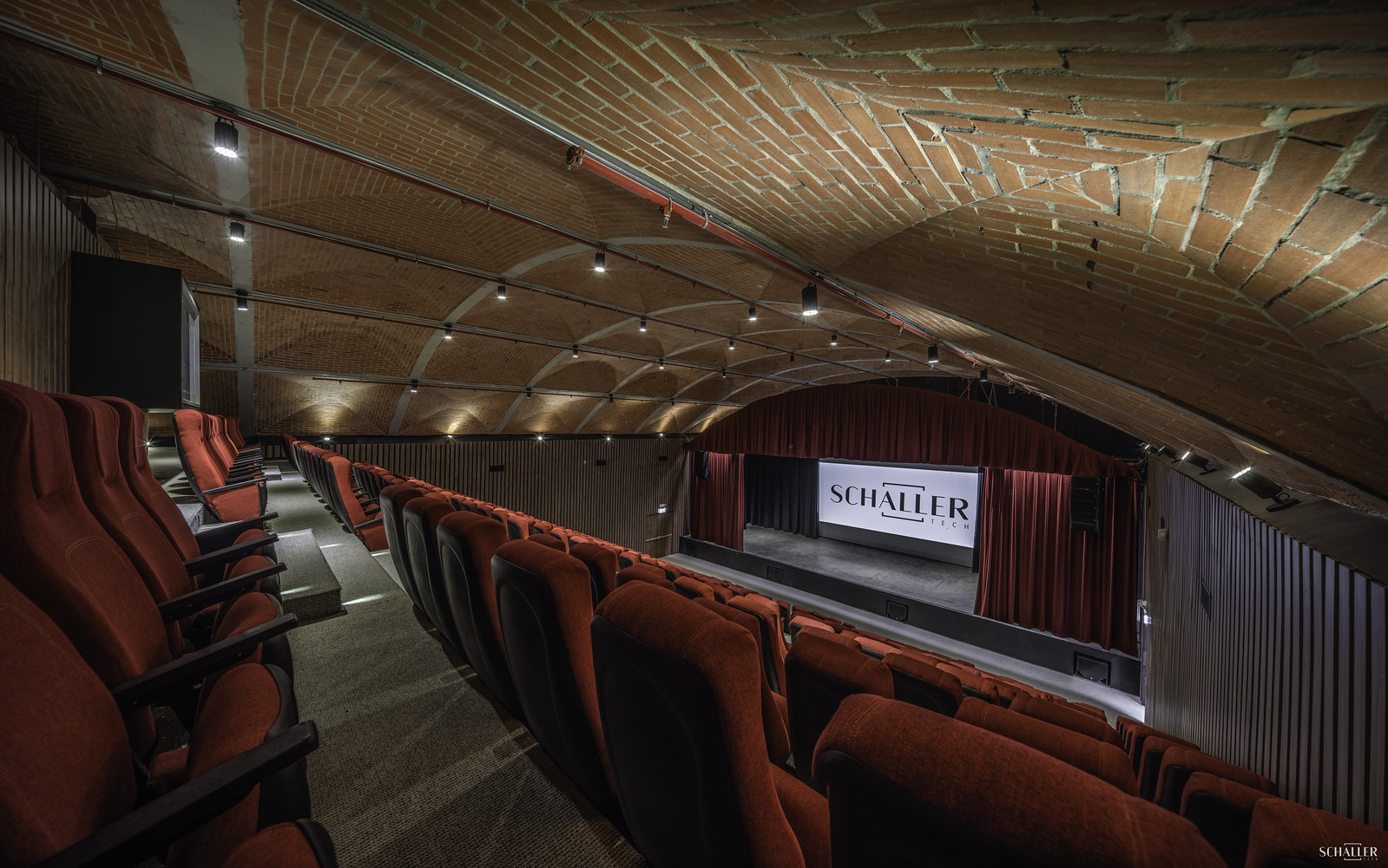 Essential AV Integration Design Fundamentals 1 An elegant modern theater interior with arched wooden ceiling, plush red seats, and a stage featuring the word ‘SCHALLER’, exemplifying ‘Essential AV Integration Design Fundamentals’. Un elegante interior de teatro moderno con techo arqueado de madera, asientos rojos de felpa y un escenario que presenta la palabra ‘SCHALLER’, ejemplificando los ‘Fundamentos esenciales del diseño de integración AV’.