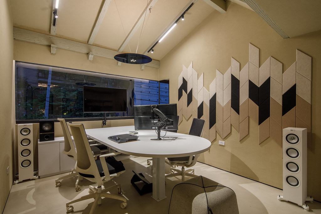 Maximizing Acoustics: The Vital Role of NRC in Sound Design 1 A modern and elegant recording or editing studio, featuring a large TV screen and built-in speakers on a beige wall, a round white table with ergonomic chairs, and a 3D geometric wall decoration with black and brown acoustic panels, optimized for an article on ‘Maximizing Acoustics: The Vital Role of NRC in Sound Design’. Un estudio de grabación o sala de edición moderna y elegante, con una gran pantalla de televisión y altavoces empotrados en una pared beige, una mesa redonda blanca con sillas ergonómicas, y una decoración de pared geométrica tridimensional con paneles acústicos negros y marrones, optimizado para un artículo sobre ‘Maximizando la Acústica: El Rol Vital del NRC en el Diseño del Sonido’.