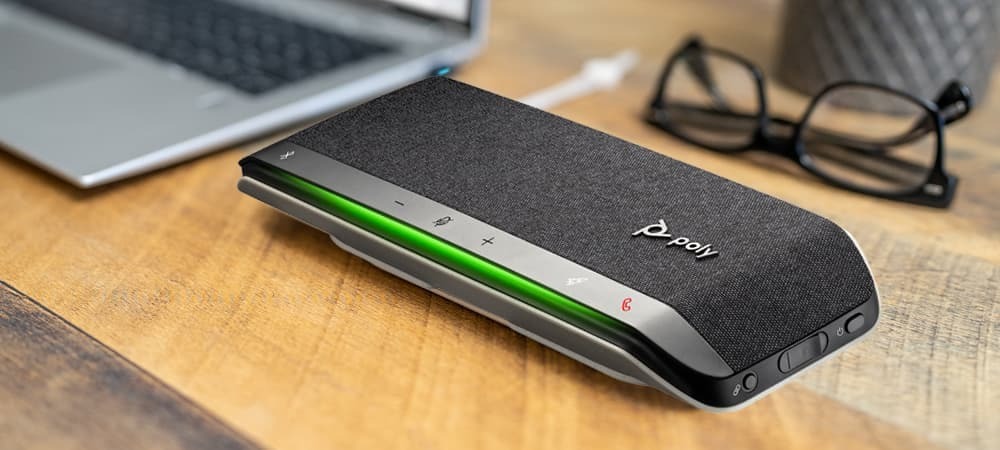 Los 5 Mejores Micrófonos para Experiencias Óptimas de Videoconferencia 3 A Poly speakerphone on a wooden surface with a laptop and eyeglasses in the background, illustrating the importance of sound quality for optimal video conferencing experiences.